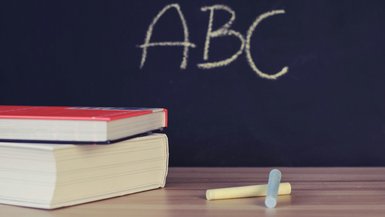 Im Vordergrund ein Tisch auf dem zwei Bücher und Kreide liegt. Im Hintergrund eine Tafel auf der ABC geschrieben steht.