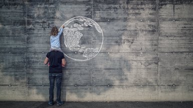 Mann hat sein Kind auf der Schulter. Diese zeichnet mit Kreide einen Globus an eine Steinwand.