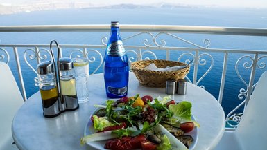 Gedeckter Tisch mit Salat und einer Flache Wasser. Im Hintergrund das Meer.