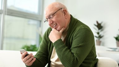 Älterer Mann mit Hörgerät blickt auf sein Smartphone und lacht.